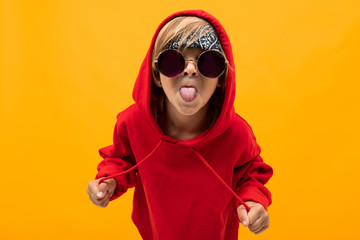 blond boy with a bandana on his head in a red hoodie with glasses shows his tongue on an orange background