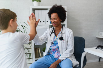Smiling female afro american doctor general practitioner talks and amuses child before medical...