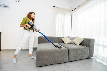 Young woman cleaning sofa with cordless vacuum cleaner in the modern white living room. Concept of...