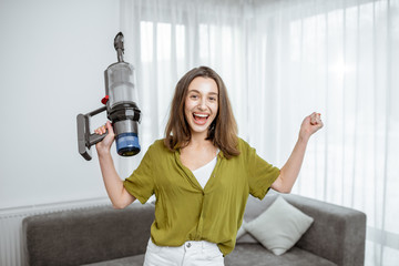 Portrait of a young and happy woman with cordless vacuum cleaner, enjoying housework at home....