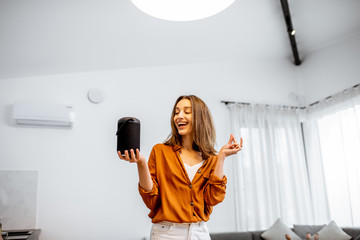 Young cheerful woman controlling home devices with a voice commands, talking to a smart column at...