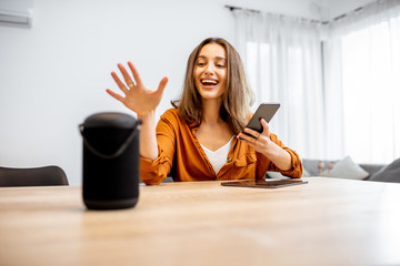 Young cheerful woman controlling home devices with a voice commands, talking to a smart column at...