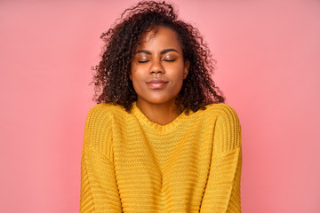 Joyful satisfied black woman with curly bushy haircut, has high self esteem, closes eyes from enjoyment, likes her new comfortable yellow sweater, stands over pink background. Love yourself concept.