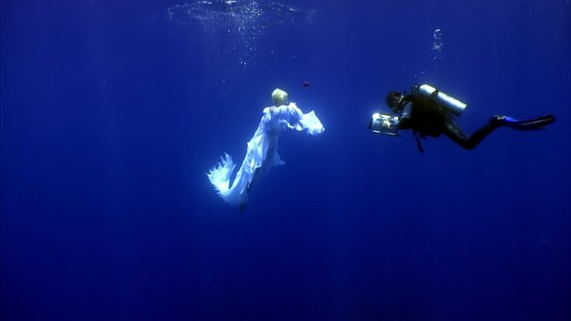 Angel young woman underwater model free diver in white cloth and cameraman. Creativity and art of creating images on background of blue water.