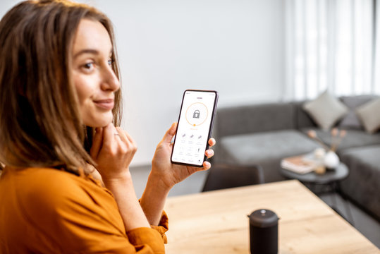 Young Woman Holding Smart Phone With Launched Security Application At Home. Concept Of Controlling And Managing Home Security From A Mobile Device