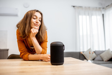 Portrait of a young and cheerful woman with a smart wireless column and phone at home. Concept of smart home and voice command control