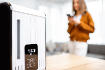 Close-up on a smart air humidifier with touch screen, woman controlling it with smart phone on the...