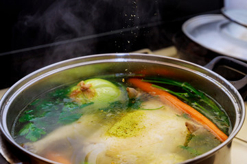 Pouring seasoning into boiling chicken broth with vegetables in steel pot.