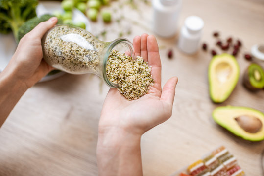 Holding Hemp Seeds At The Table With Healthy Vegan Food Ingredients And Supplements, Top View. Concept Of Marijuana As A New Trend In Food And Cosmetic Industry