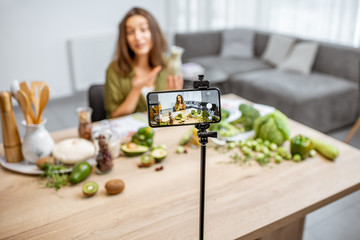 Young woman recording on a smart phone her vlog about healthy eating. Sitting at the table with lots of green vegan food ingredients at home
