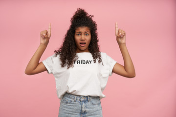 Confused attractive dark skinned curly brunette woman with casual hairstyle frowning eyebrows and looking at camera with pout, standing over pink background with raised forefingers