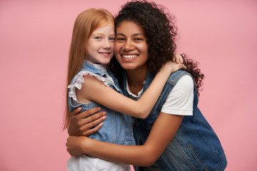 Couple of happy lovely young ladies in jeans vests and white shirts embracing each other lovingly and looking cheerfully to camera with charming smiles, posing against pink background