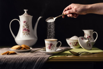 Hand of a person pouring sugar with a spoon in a hot coffee served on a vintage porcelain tableware with floral ornaments and cookies on tablecloths on a rustic wooden table. Traditional lifestyle