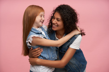 Indoor shot of two cheerful attractive young girls looking lovingly on each other and smiling sincerely, giving gentle hugs and having nice time together, isolated over pink background