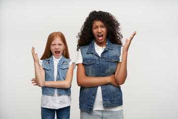 Studio shot of irritated young couple of curly dark haired female and foxy long haired girl in casual clothes looking angrily to camera and shouting annoyed, isolated over white background