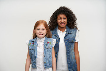 Couple of young confused pretty long haired girls wearing family look while posing over white background with hands down, grimacing their faces and frowning eyebrows