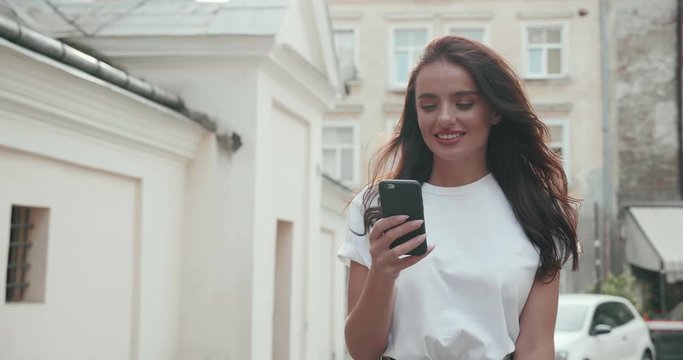 Young Pretty Woman walking in the Town on sunny day, while using her Mobile Phone. Having online Conversation. Looking Happy and Satisfied with her Outfit of the Day. Having charming smile