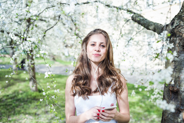 Happy young woman holding the phone and listening to music or podcast via earphones in a white t-shirt under the blooming cherry trees. Smiling, looking at the camera.