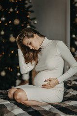 Pregnant woman sits on bed against background of Christmas tree.