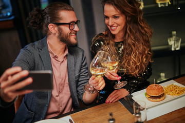 Cute caucasian fashionable couple sitting in restaurant at dinner holding glasses of wine and taking selfie.