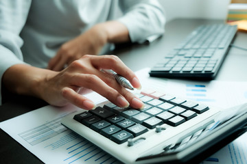 The hand of Asian woman is pressing calculator and prepare information for report to correct. While still holding a silver pen in finger. When calculated completed, can be recorded immediately.