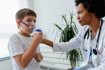 Woman African American doctor general practitioner helping child to put nebulizer inhaler face...