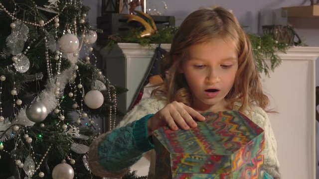 Surprised blond 8 years old girl opens present box next to the Christmas tree