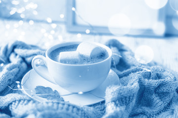 Obraz na płótnie Canvas Cup of coffee with marshmallow, knitted scarf, garland. Hygge concept. Soft focus. Toned classic blue trend color