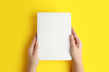 Woman holding book with blank cover on yellow background, top view