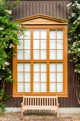 Window with curtains and shutters on a wooden house from boards with flowers and a bench