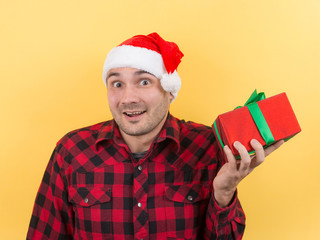 Happy surprised man in a red hat holding a gift with admiration on his face. Positive human emotions at christmas. Yellow background, copy space.