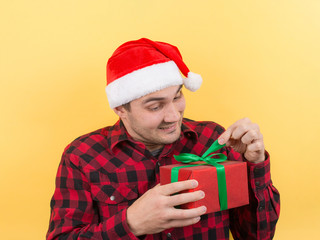 Pensive man in a red hat, holds a gift with interest on his face. Positive human emotions for christmas, secret santa. Yellow background, copy space.