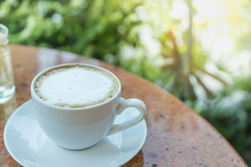 Close up of white cup and plate of hot latte coffee on round table and green leaf nature background.