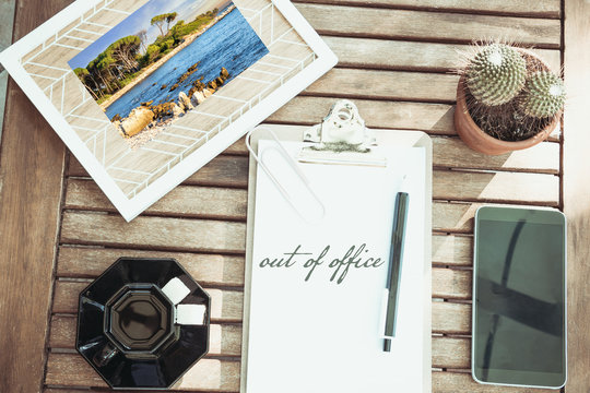 Bureau en flat lay avec une photo de vacances un café un smartphone et un cactus les mots Out of office écrits en anglais sur un bloc-note avec son stylo bureau en bois