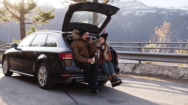Loving, young couple drinks hot tea from thermos flask sitting in car trunk. Happy caucasian couple having a coffee break during road trip in countryside - drink and eating sandwiches. Admiring the