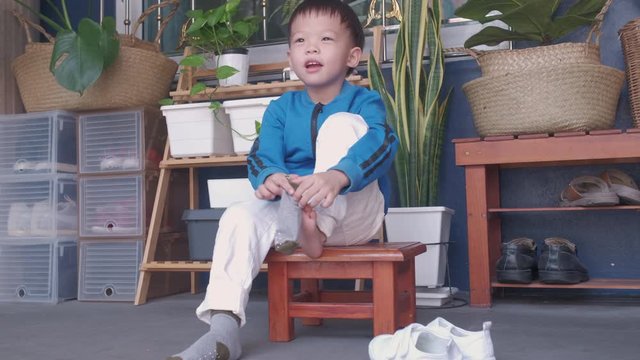 Cute little Asian 3 years old toddler kindergarten kid sitting near shoe rack near front door of his house and concentrate on putting on his own socks, Encourage Self-Help Skills in Children concept
