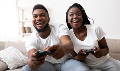 Excited afro couple playing video games at home together using joysticks