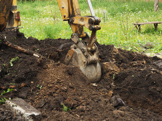 An excavator digs a hole in the ground. Summer, grass, dandelions, a wooden bench and an apartment building.. © Olga