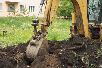 An excavator digs a hole in the ground. Summer, grass, dandelions, a wooden bench and an apartment building..