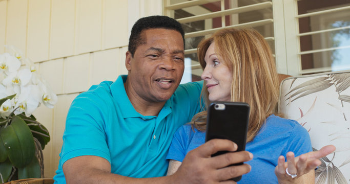 Smiling older couple sitting on porch and using cell phone and laughing together