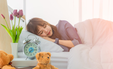 Alarm clock on bedside table with flower vase and bear doll by young woman sleeping with happy in morning is background.