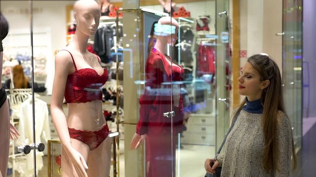 A beautiful slender young Shopaholic woman walks up to the window of a lingerie and homewear store in the Mall and looks at a mannequin in luxurious red lingerie. Purchases. Sale.