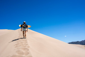 woman with sand board walking on edge of a sand dune in the great sand dunes national park,...