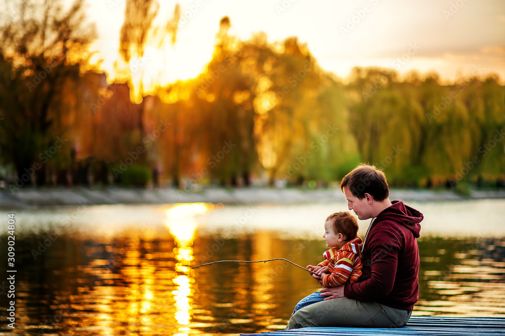 Wall mural Father and 2 years old child play fishing on bank of lake in sunset time. Golden in sun light trees and water around. Father and daughter son fun together. Lesson of fishing in evening time. Weekend