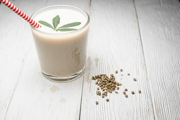 Hemp milk is poured into a glass. On the surface is a cannabis leaf. Nearby marijuana seeds.