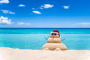 Fototapeta na wymiar Smiling sandy snowman with red santa hat on the caribbean beach with beautiful sky . Holiday concept for New Year and Christmas Cards