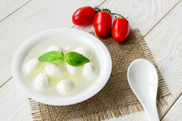 Mini mozzarella cheese balls with brine, three red cherry tomatoes and porcelain spoon on a  white rustic wooden table. Healhy mediterranean cuisine and vegetarian food.