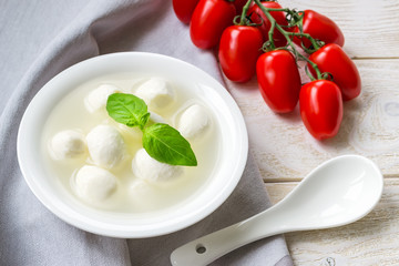 Mini mozzarella cheese balls with brine, branch of red cherry tomatoes and porcelain spoon on a  white rustic wooden table. Healhy mediterranean cuisine and vegetarian food.