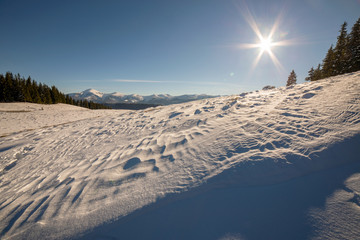 Winter landscape panorama with snowy landscape hills, distant white mountains, dark forest and clear blue sky with bright sun.