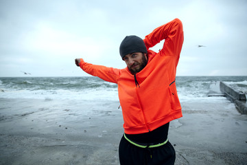 Young handsome sporty dark haired bearded man doing stretching exercises outdoor, preparing for morning workout, dressed in black warm athletic clothes and orange coat with hood